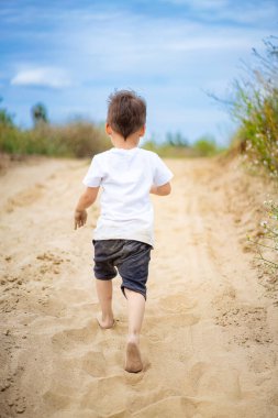Çocuk gündüz vakti kumlu yolda. Genç bir çocuk açık bir gökyüzünün altında çimlerle ve kır çiçekleriyle çevrili kumlu bir patikada yalınayak koşuyor..