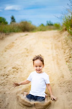 Çocuk kumlu bölgede neşeyle oynuyor. Küçük bir çocuk kumlu bir alanda oturur, gülümser ve oynar, doğayla çevrili güneşli bir günün tadını çıkarır..