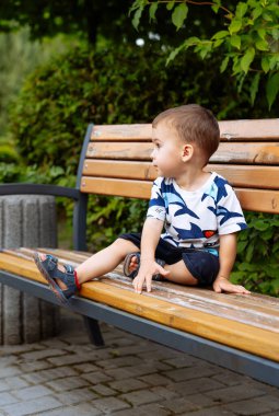 Parktaki bankta oturan çocuk. Günlük giysiler içindeki küçük bir çocuk, sakin bir anın tadını çıkarırken bir bankta oturur ve yan tarafa bakar..