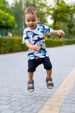 Küçük çocuk neşeyle parkta zıplıyor. Parkta oynayan genç bir çocuk, güneşli bir günde çevresi yeşilliklerle çevrili bir yolda neşeyle zıplıyor..