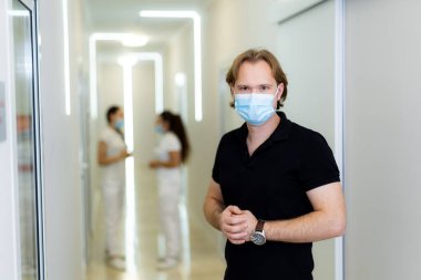 Modern bir klinikteki doktor. Maske takan bir adam koridorda dikilirken, arka planda iki kadın konuşarak sağlık hizmetlerini ön plana çıkarıyor..