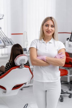 Hasta bakımına hazırlanıyorum. Diş hekimi eldiven takan bir uzman klinikte güvenle ayakta dururken hasta diş hekimliği koltuğunda oturuyor..