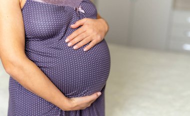 Hamile anne karnı tok. Benekli elbiseli hamile bir kadın hafifçe aydınlatılmış bir odada karnını nazikçe tutuyor..