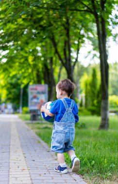 Park yerinde futbol topu olan bir çocuk. Tulumlu küçük bir çocuk ağaçlar ve yeşil çimlerle çevrili bir futbol topunu taşıyan bir patika boyunca yürür..