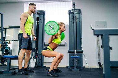 Spor salonunda birlikte birkaç egzersiz. Gündüz vakti spor salonunda ağırlıklarla çömelirken bir erkek bir kadına koçluk yapıyor..