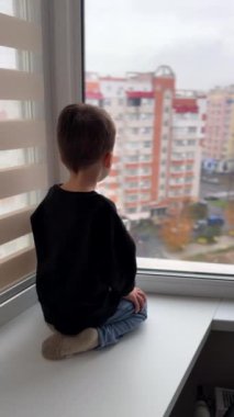 Pencere pervazında oturan bebeğin arka manzarası. Çocuk caddelerde hareket eden apartmanlara ve arabalara bakıyor. Dikey video.