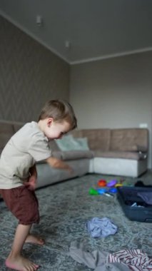 Neşeli beyaz çocuk kıyafetlerini tavana fırlatıyor. Arka planda bir bavul ve yere saçılmış oyuncaklar. Dikey video.