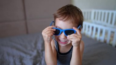 Sevimli gülümseyen beyaz bebek güneş gözlüğünü takıyor. Aktif çocuk ayağa kalkar ve neşeyle büyük yatağa atlar..