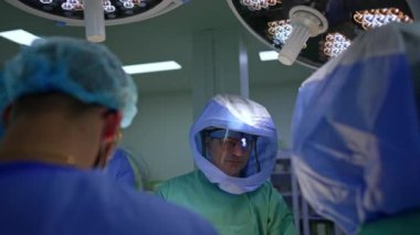 Farlı, koruyucu başlıklı ve bornozlu bir erkek doktor bir süre doğrudan kameraya bakar. Travma ameliyatı yapan bir takımda çalışan bir cerrahın portresi..