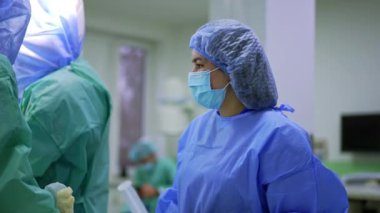 Ameliyat odasında kep, maske ve üniforma giyen kadın ameliyat hemşiresi duruyor. Cerrahların taleplerini bekleyen doktorlara yardım.
