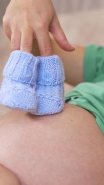 Hamile bir kadın ve gelecekteki çocuk için küçük çoraplar tutuyor. Hamile bir kadının göbeğinin yan görünüşü, örgü örülmüş bebek çorapları. Dikey video