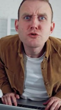 Evde çalışan komik komedyen. Mutfak masasında dizüstü bilgisayarla otururken kızgın bir şekilde kameraya bakan genç bir serbest çalışanın kızgın yüzü. Dikey video