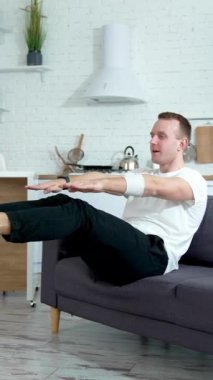 Genç adam evde antrenman yapıyor. Yakışıklı adam kanepede oturuyor ve mutfakta karantina sırasında egzersiz yapıyor. Dikey video