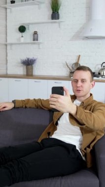 Genç adam web kamerası kullanıyor. Adam kanepede otururken video çekiyor. Yakışıklı bir erkek blogcu dairesinden görüntülü konuşma yapıyor. Dikey video