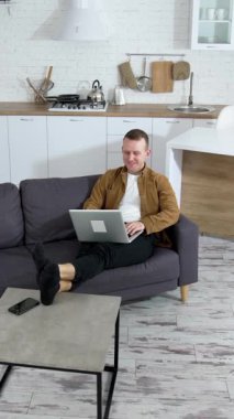 Dizüstü bilgisayarda çalışan genç bir iş adamı. Modern mutfak geçmişi ve bir kanepede dizüstü bilgisayarla oturan bir adam. Uzaktan kumanda. Dikey video