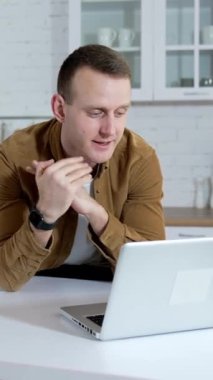 Genç adam mutfak arka planında görüntülü konuşma yapıyor. Freelancer evdeki kablosuz bilgisayarda çalışıyor. Uzaktan kumanda. Dikey video