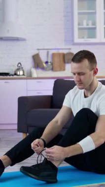 Eğitimden önce genç adam ayakkabı bağını evde bağlıyor. Paspasın üzerinde oturan ve spor yapmak için siyah spor ayakkabılarını giydiren adam. Dikey video