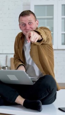 Evde oyun oynayan duygusal genç adam. Mutfak masasında dizüstü bilgisayarla otururken kamerayı işaret eden komik adam. Dikey video