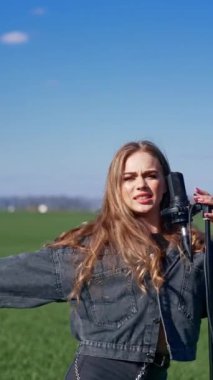 Yeşil alanda performans sergileyen çekici bir kadın. Kot elbiseli güzel bir kız mikrofonda şarkı söylüyor ve dışarıdaki kameraya bakıyor. Dikey video