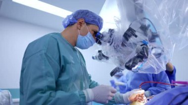 Mikroskoba bakan beyin cerrahı sol eliyle elektrikli alet kullanıyor. Sağlık personeli arka planda çalışıyor..