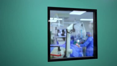 Ameliyat odasına kare pencereli mavi kapılar. Doktor ameliyathanede mikroskop kullanarak ameliyat yapıyor..