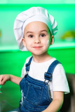 Genç aşçı fırın eğlencesine hazırlanıyor. Aşçı şapkalı çocuk yemek pişirme aletleriyle dolu renkli bir mutfakta pişirmeye hazırlanırken ışıl ışıl gülümsüyor..