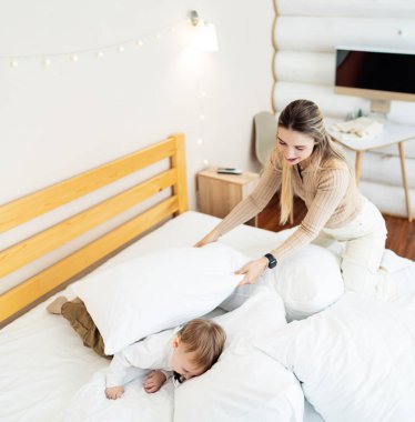 Sıcak yatak odasında oyuncu anne ve çocuk. Yavrusu modern, rahat bir yatak odasında onların altında saklanırken bir anne neşe içinde yastıkları düzenliyor..