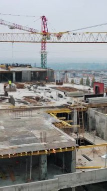 Alışveriş merkezinin yeni şantiyesi. Şehrin arka planında inşaat sırasında çatıda işçiler ve büyük vinçlerle hava manzarası. Drone geri gidiyor. Dikey video