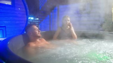 İnsanlar jakuzide oturuyor ve üstlerinde bir sürü buhar yükseliyor. Çizgili mayo giymiş kadın telefonda konuşuyor..