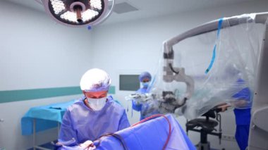 Profesyonel beyin cerrahı ameliyat yapan hastanın başında oturur. Kablolu modern cihazlar hastanın üzerinde. Sıhhiye mikroskobu plastikle kaplıyor.