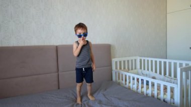 Güneş gözlüğü takan mutlu çocuk çift kişilik yatakta zıplıyor. Bebek evde, yatak odasında eğleniyor..