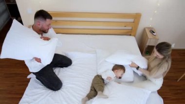 Sevgi dolu ebeveynler yataklarında oturup küçük bebeklerine neşeyle vuruyorlar. Ağzında emzik olan çocuk gülüyor ve oyun oynamayı seviyor. Üst görünüm.