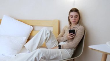 Sarışın kadın sandalyeye oturup bacaklarını yatağa koyuyor. Bayan akıllı telefonunu kullanıyor..