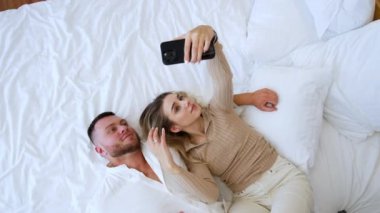 Bir çift yatakta uzanıp sarılıyor. Gülümseyen sarışın kadın elinde telefonla selfie çekiyor. Üst görünüm.