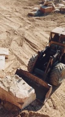 Profesyonel ekipman bir kovayla bir taşı devirip arka plandaki kumlu taş ocağının yakınına kaldırıyor. Yakın plan. Dikey video