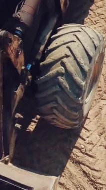 Kocaman tekerlekleri ve kovası olan buldozer kumlu bir taş ocağının bölgesinde hareket ediyor. Yakın plan. Dikey video