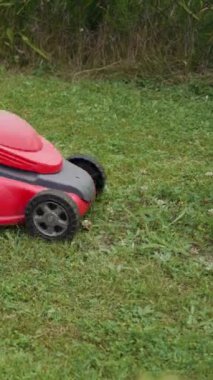 Bahçede çim biçme makinesiyle çim biçen çıplak ayaklı genç bir kız. Çıplak kadın ayakları arka bahçede çim biçme makinesinin arkasında yürüyor. Dikey video