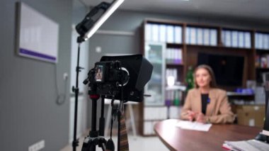 Tripodun üzerinde görüntüsü olan profesyonel kamera bir video kaydediyor. Bej ceketli kadın el kol hareketi yapıyor. Bulanık arkaplan.