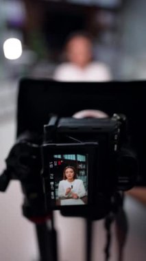 Esmer, uzun saçlı bir kadını kaydeden profesyonel bir kamera görüntüsünden uzaklaşmak. Günlük oluşturma. Arkaplan bulanık. Dikey video.