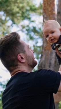 Sakallı beyaz bir adam yeni doğmuş şirin bir çocuğu kafasından tutuyor. Mutlu baba bebeğine hayranlıkla bakıyor. Düşük açı. Arka planda çam ağaçları. Dikey video