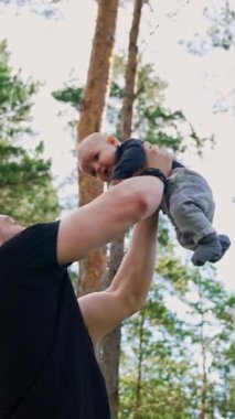Yakışıklı, kaslı, beyaz bir adam bebeğini fırlatıyor. Doğada çocukla vakit geçirmek. Düşük açı. Arka planda çam ağaçları. Dikey video