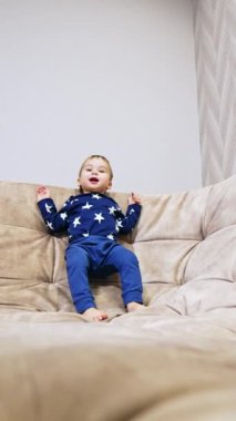 Beyaz tatlı çocuk büyük kanepede zıplıyor. Küçük oğlan gülümsüyor ve mutlu bir şekilde el sallıyor. Dikey video