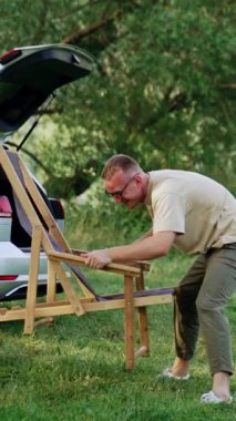 Orta yaşlı, beyaz erkek, çayırdaki sandalyeyi açıyor. Adam ormanda dinlenmek için hazırlanıyor. Dikey video