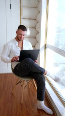 Bacağında dizüstü bilgisayar tutan bir adam. Evde çalışan mutlu, rahat bir insan. Dikey video.