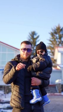 Sakallı adam ve oğlu kalın ceketler ve güneş gözlüğü takıyor. Babası kameraya görünür ama çocuk yere düşmek ister. Dikey video.