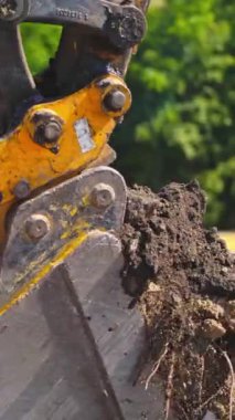 Bir kova traktör zemini kazıyor. Kazıcı yazın yeşil ağaçların arka planındaki toprağı kazıyor. Yakın plan. Dikey video