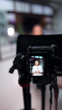 Çekici beyaz kadın kameranın önünde tripodla konuşuyor. Kapatın. Blog için video içeriği oluşturma. Arkaplan bulanık. Dikey video.