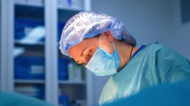 Mavi şapkalı, maskeli ve cüppeli kadın doktor. Ameliyat sırasında hastanın üzerine eğilen bir cerrahın portresi..
