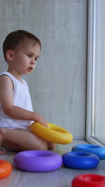 Beyaz takım elbiseli tatlı erkek bebek elinde oyuncakla yerde oturuyor. Ciddi bir çocuk pencereye bakar ve bir şeyler söyler. Dikey video