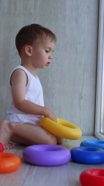 Sevimli beyaz erkek bebek balkonda oynuyor. İlk çocuk pencereye bakar ve piramit toplar. Dikey video